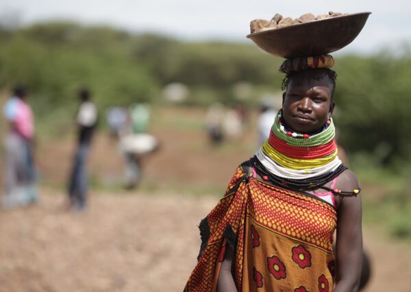 Frau in traditionellem sudanesischen Gewand und vielen bunten Ketten um den Hals balanciert eine Schale mit Steinen auf dem Kopf mit denen sie ihr Einkommen sichert.