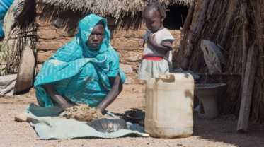 Eine Frau kniet in traditioneller Kleidung neben einem kleinen Kind vor einer Hütte in einem ländlichen Gebiet im Sudan. Vor ihnen steht ein Kanister, sie knetet eine Masse auf einem Stück Stoff.